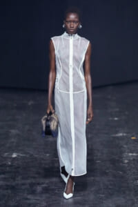 Model walks the runway in a sheer white sleeveless gown with a central zipper, carrying a fur-trimmed handbag and white heels.
