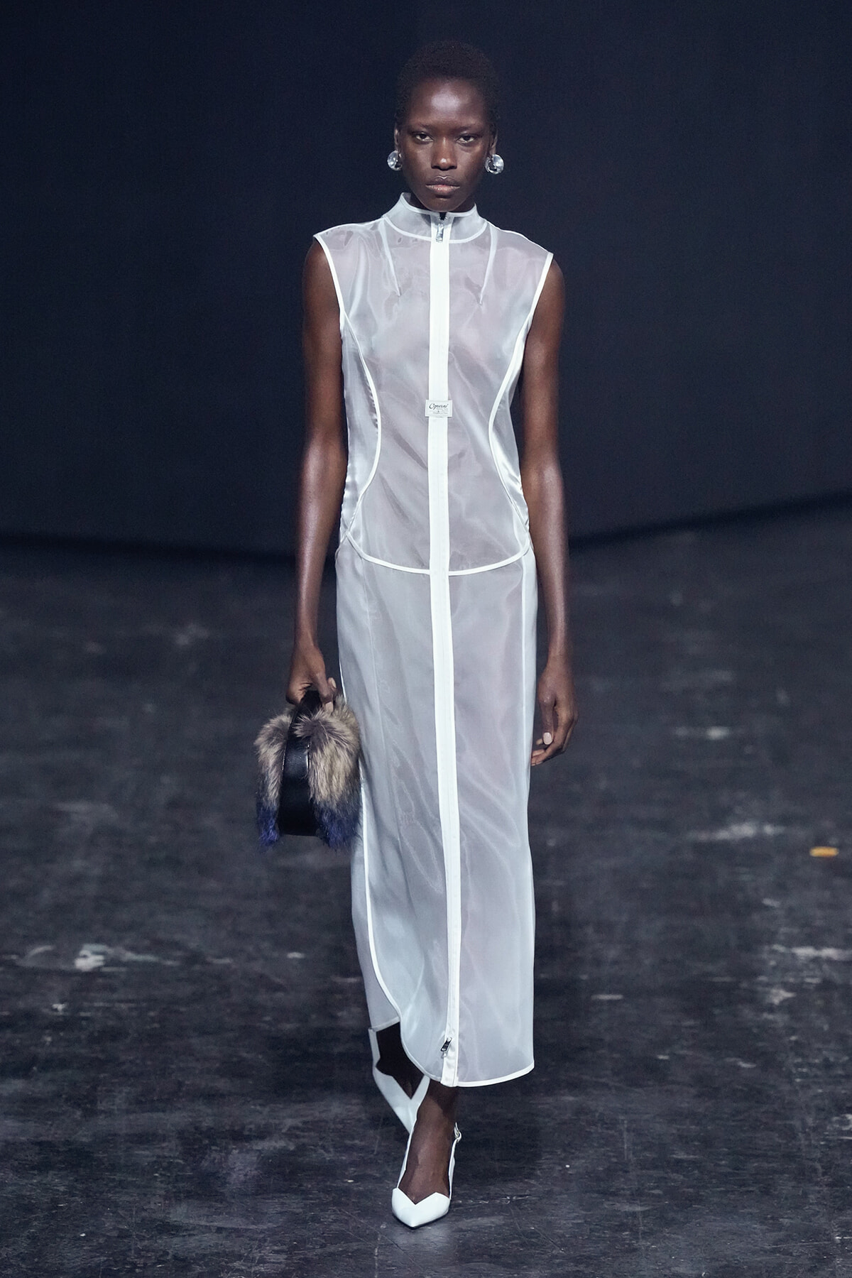 Model walks the runway in a sheer white sleeveless gown with a central zipper, carrying a fur-trimmed handbag and white heels.