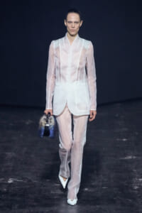 Model walks runway in sheer white suit with matching trousers, holding a blue-gray fur handbag before a dark backdrop.