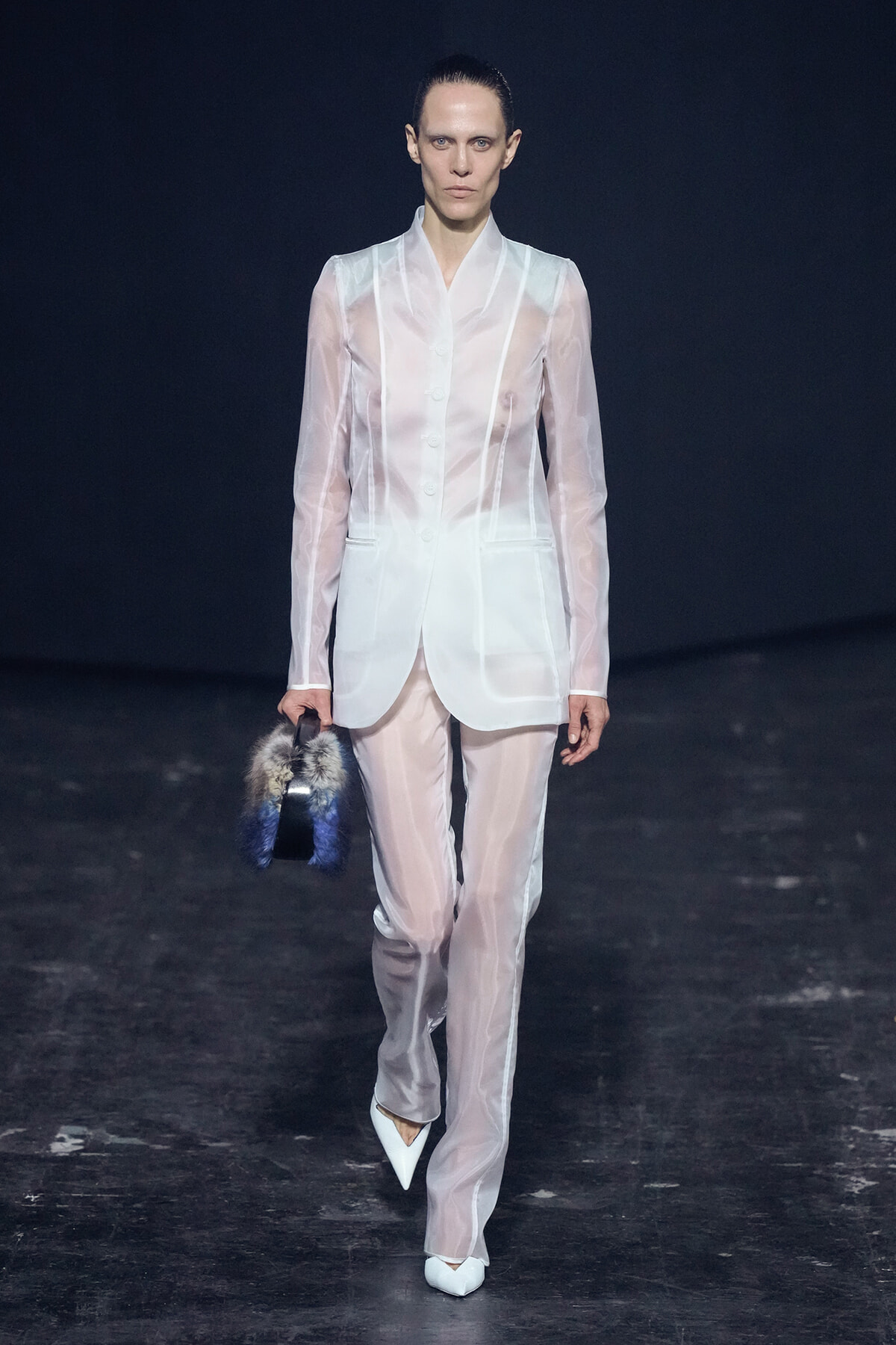 Model walks runway in sheer white suit with matching trousers, holding a blue-gray fur handbag before a dark backdrop.
