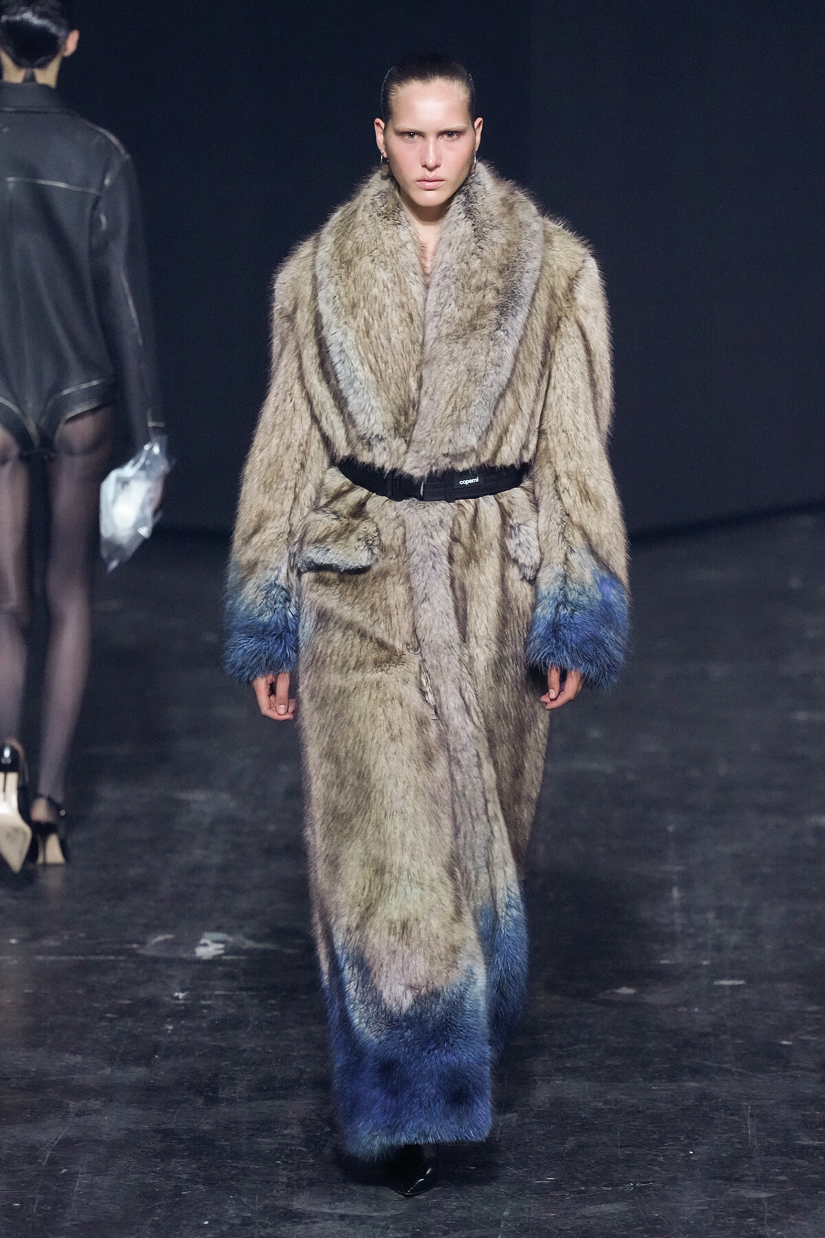 Female model walks a fashion runway wearing a long beige fur coat with blue gradient cuffs and a black belt.