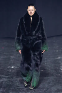 Model on a runway wearing a long black fur coat with green-fade cuffs and a waist belt, walking forward.