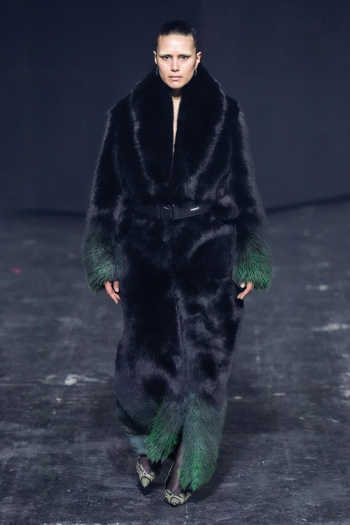 Model on a runway wearing a long black fur coat with green-fade cuffs and a waist belt, walking forward.