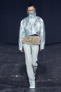 Model on runway wearing a metallic silver high-collar jacket with a fur belt and light gray pants.