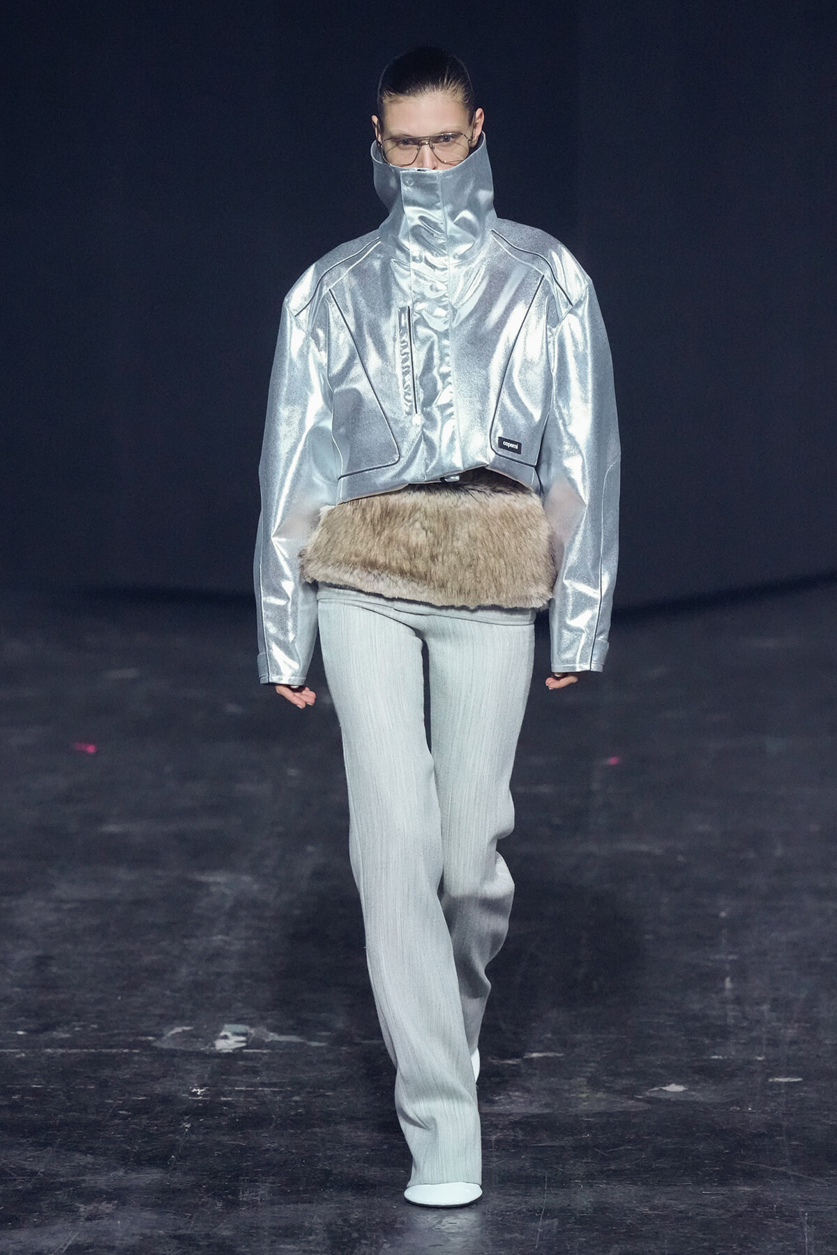 Model on runway wearing a metallic silver high-collar jacket with a fur belt and light gray pants.