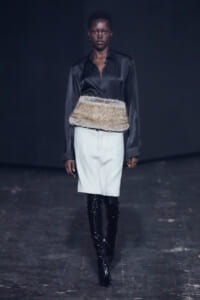 Model walks the runway in a black satin blouse tucked into a light pencil skirt, accented by a wide fur belt and shiny black knee-high boots.