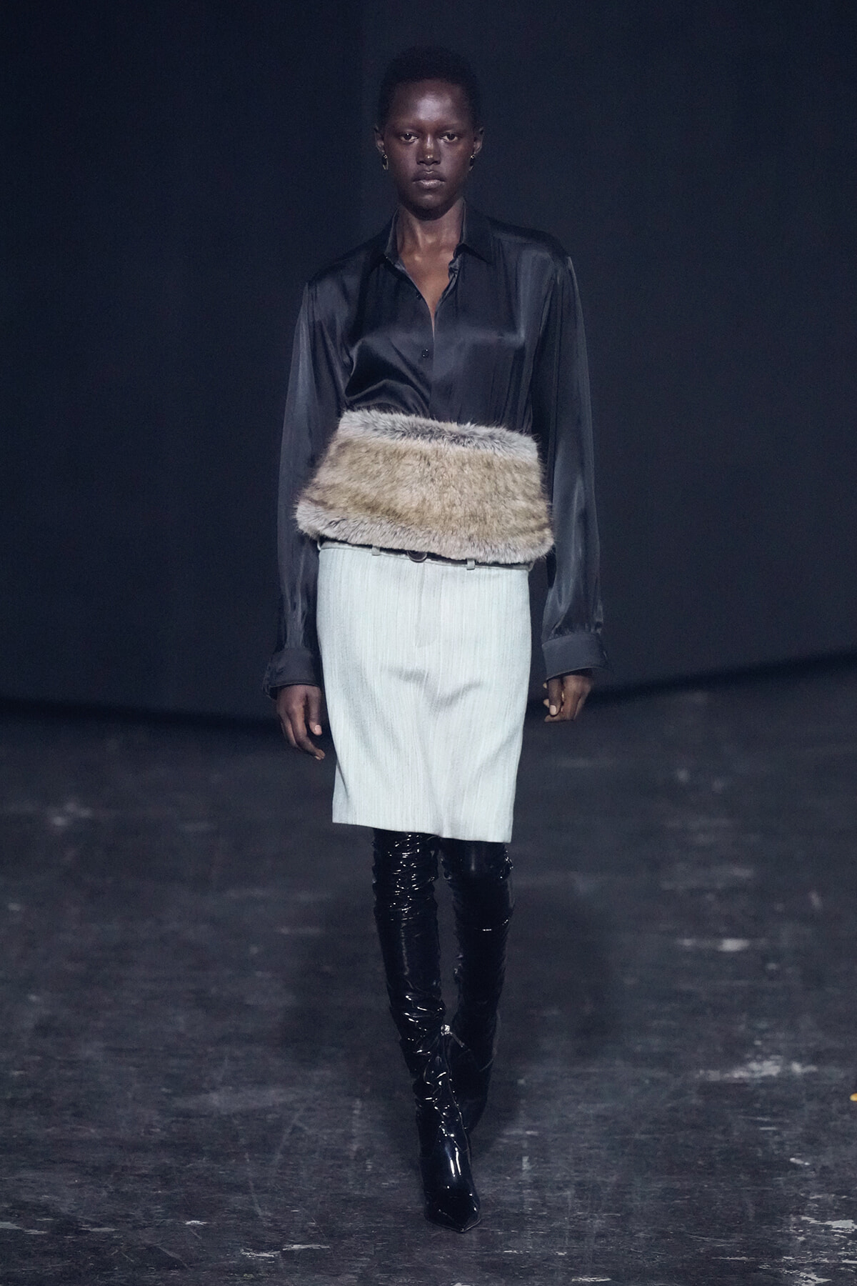 Model walks the runway in a black satin blouse tucked into a light pencil skirt, accented by a wide fur belt and shiny black knee-high boots.