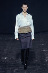 Model on runway wearing a white satin blouse tucked into a dark denim pencil skirt with a fur belt and black knee-high boots.