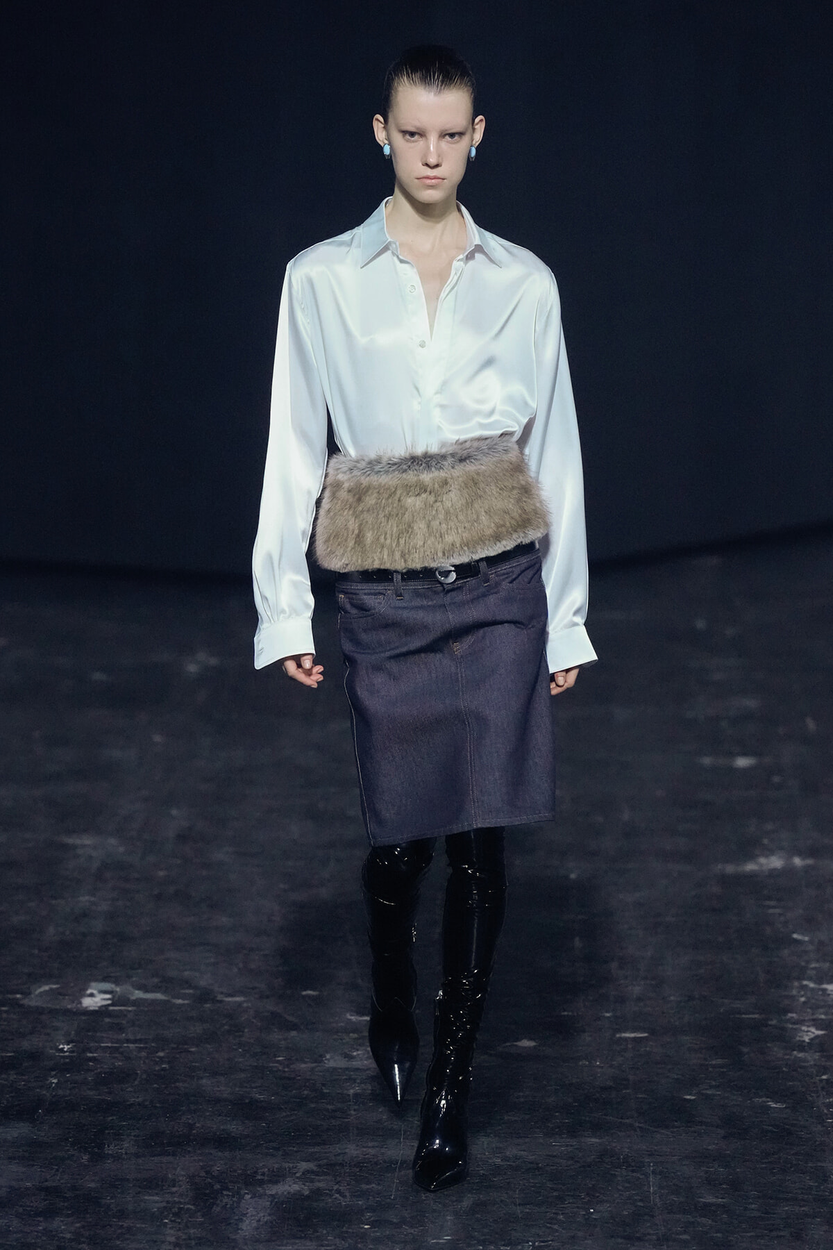 Model on runway wearing a white satin blouse tucked into a dark denim pencil skirt with a fur belt and black knee-high boots.