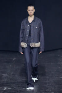 Model on a dark runway wearing a dark denim jacket and matching jeans, with two fur pompoms at the waist and white star-shaped shoes.