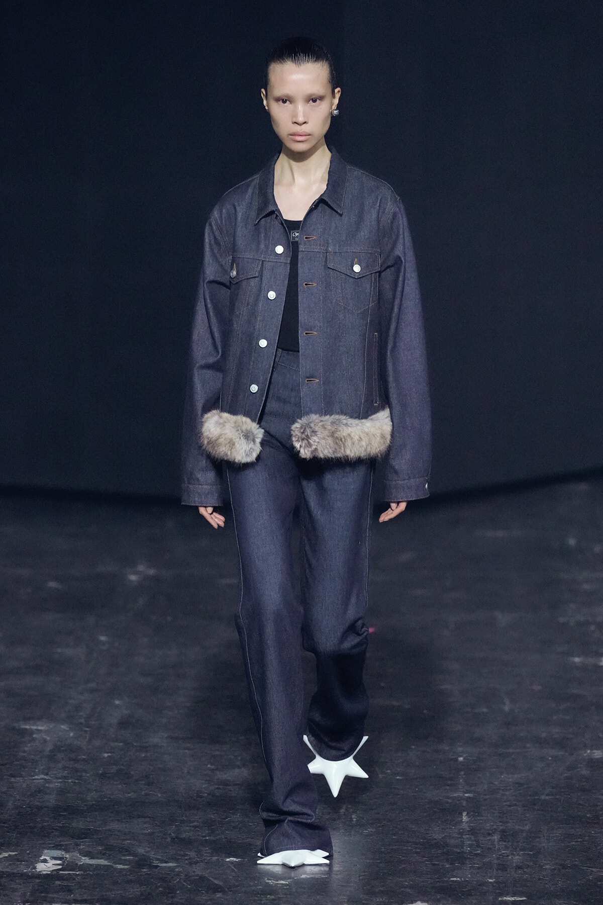 Model on a dark runway wearing a dark denim jacket and matching jeans, with two fur pompoms at the waist and white star-shaped shoes.