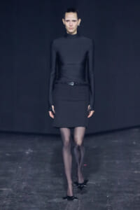 Model walks a runway in an all-black ensemble: turtleneck top, belt, knee-length skirt, sheer tights, and star-shaped heels against a dark backdrop.