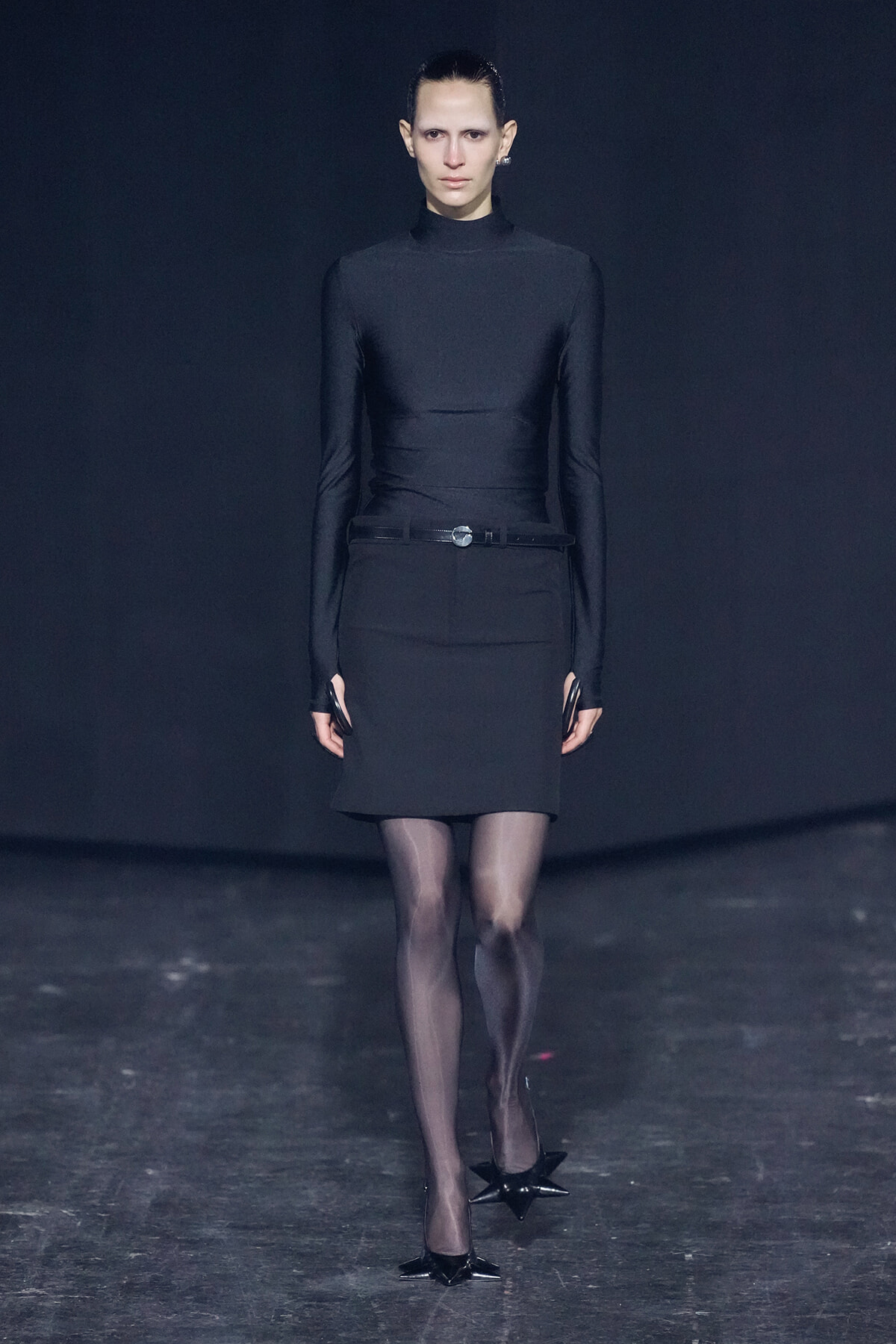 Model walks a runway in an all-black ensemble: turtleneck top, belt, knee-length skirt, sheer tights, and star-shaped heels against a dark backdrop.