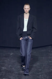 Model walking the runway in a black blazer, white top, loose dark jeans, and black pointed heels against a dark backdrop.