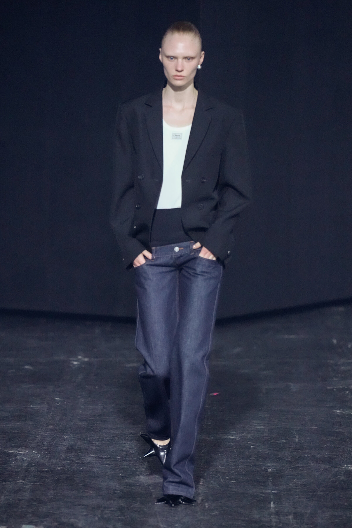 Model walking the runway in a black blazer, white top, loose dark jeans, and black pointed heels against a dark backdrop.
