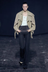 Model on a dark runway wearing a beige trench coat over a white top and black high-waisted pants with black pointed-heel shoes with star accents.