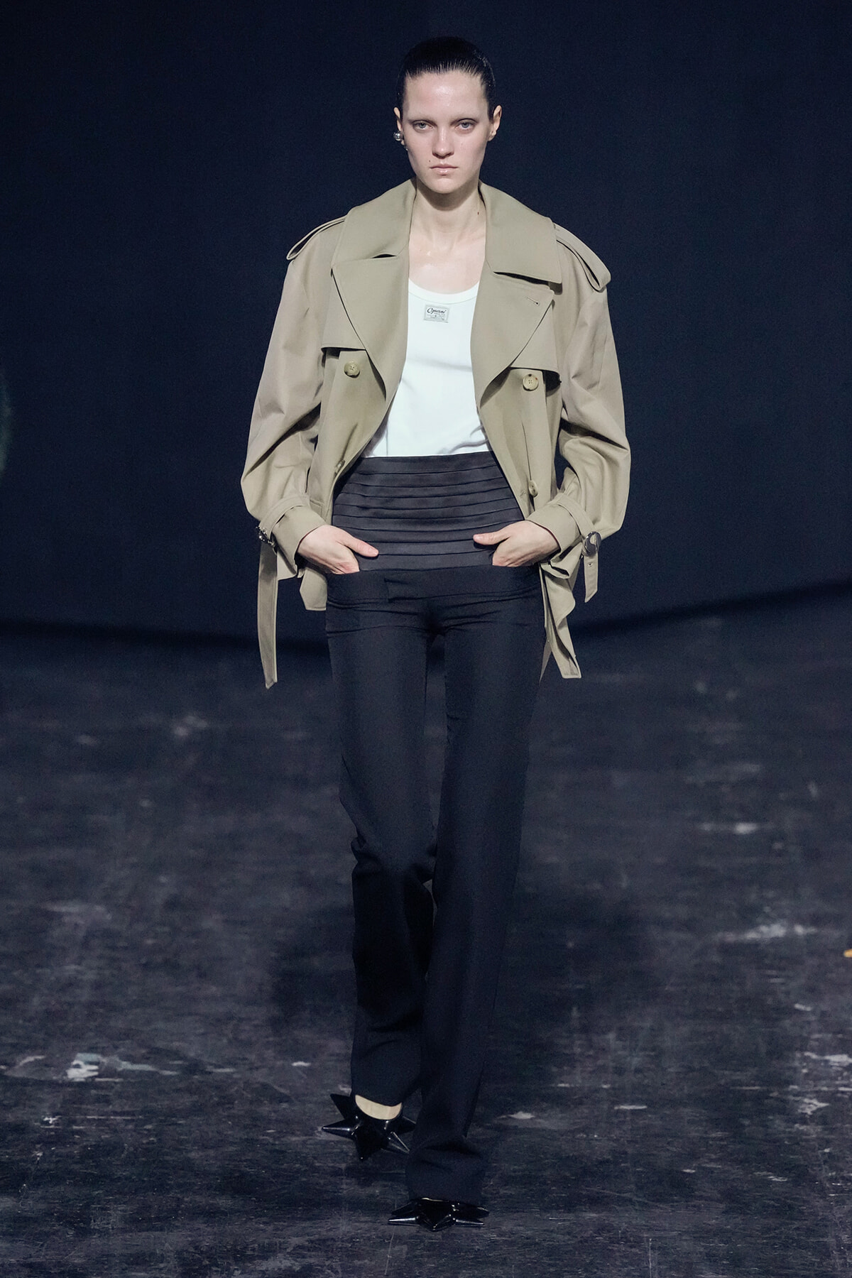 Model on a dark runway wearing a beige trench coat over a white top and black high-waisted pants with black pointed-heel shoes with star accents.