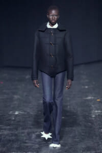 Model on a runway wearing a black toggle-coated coat, white shirt, blue jeans, and white star-shaped shoes against a dark backdrop.