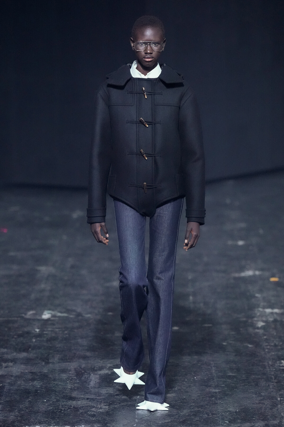 Model on a runway wearing a black toggle-coated coat, white shirt, blue jeans, and white star-shaped shoes against a dark backdrop.