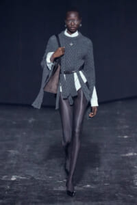 Model walking a runway in a dark setting wearing a chunky gray knit cape over a white shirt and brown belt, carrying a brown shoulder bag.