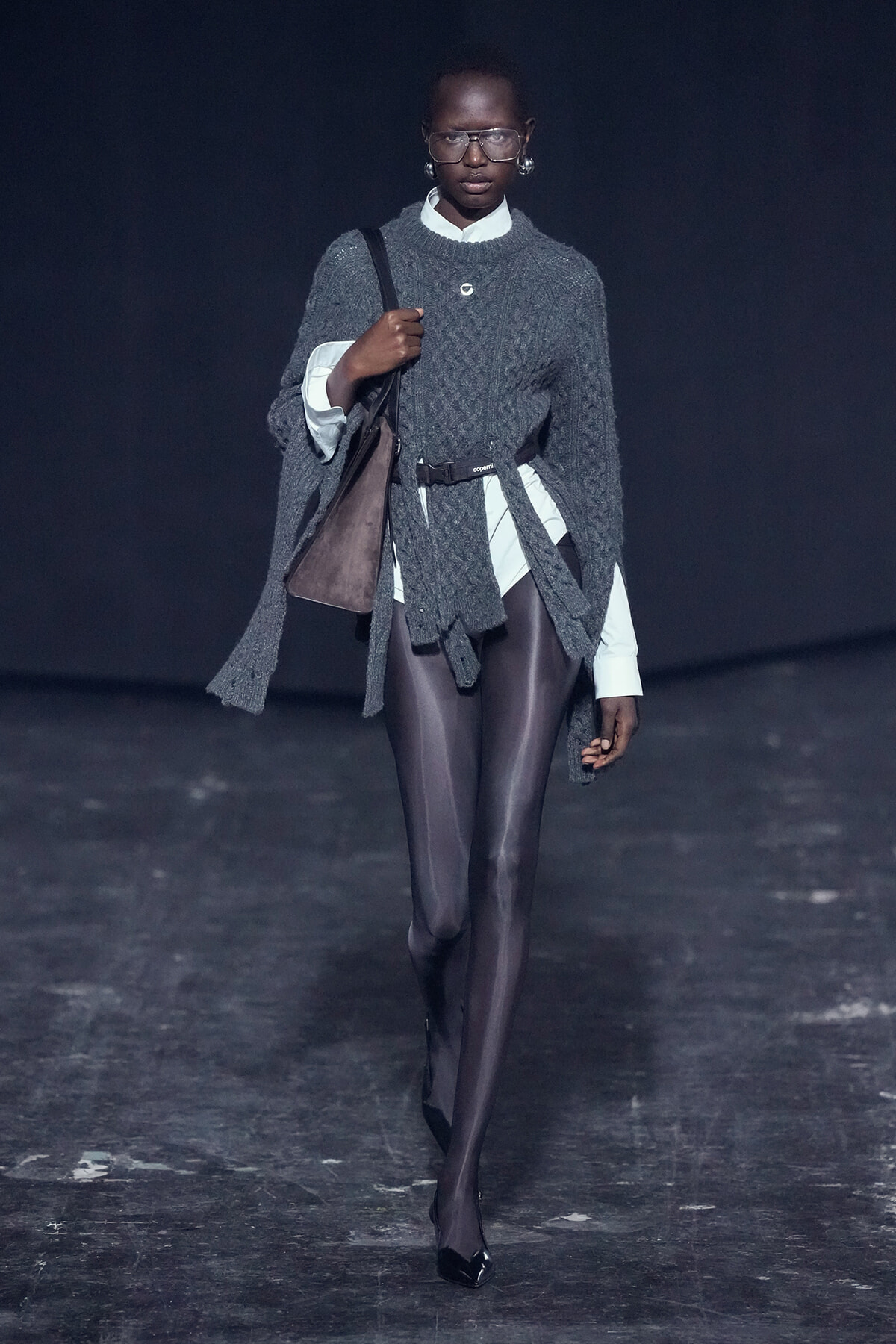 Model walking a runway in a dark setting wearing a chunky gray knit cape over a white shirt and brown belt, carrying a brown shoulder bag.