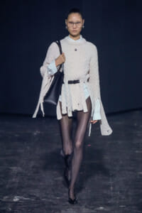Model walks runway in an oversized cream cable-knit sweater layered over a light shirt, with black tights and pointed heels, carrying a black shoulder bag.