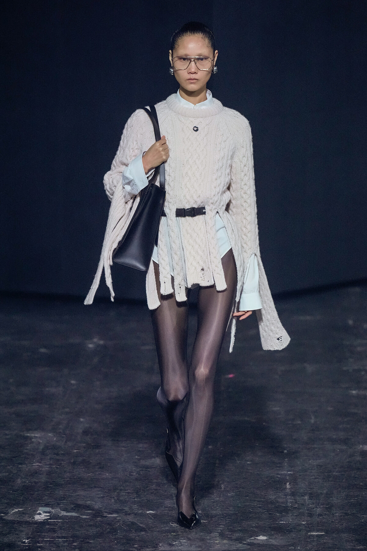 Model walks runway in an oversized cream cable-knit sweater layered over a light shirt, with black tights and pointed heels, carrying a black shoulder bag.