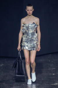 Model walking a runway in a strapless dress made of crumpled aluminum foil pieces, white sneakers, and carrying a large black tote bag.