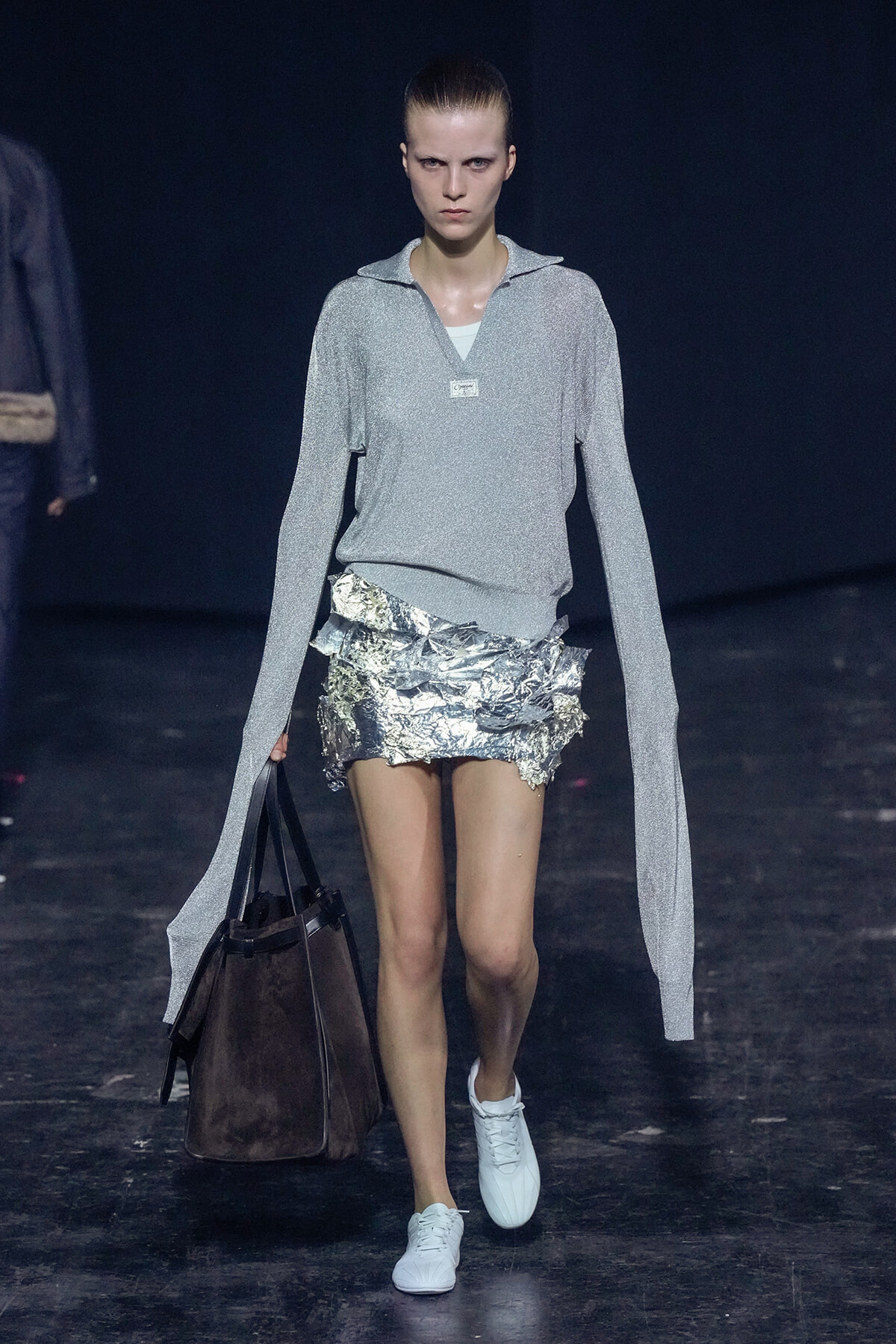 Model on a runway wearing a shimmering silver long-sleeve top and a crinkled metallic foil mini skirt, carrying a large brown tote bag.