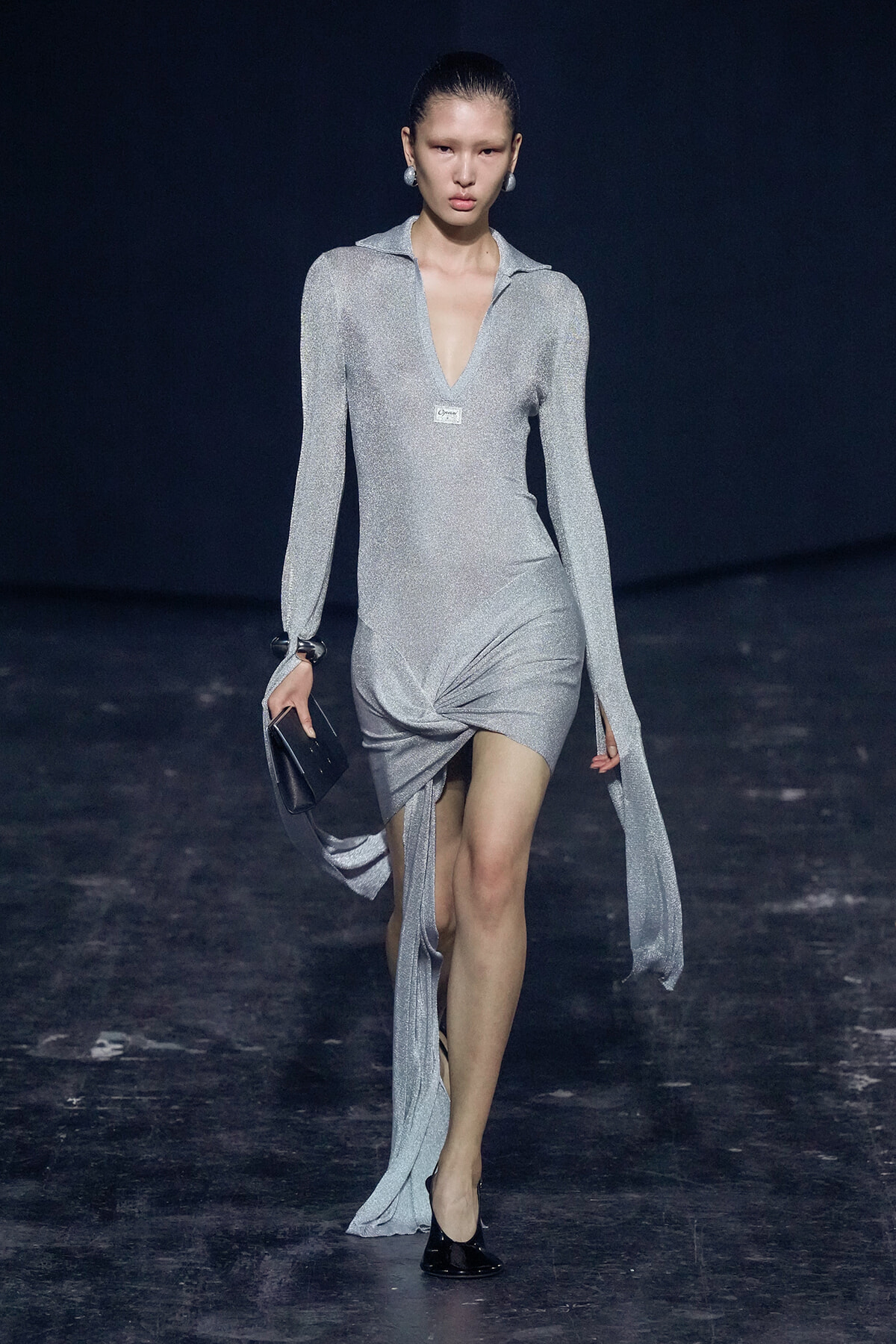 Model walks the runway in a silver glittery long-sleeve dress with a plunging V neckline and twisted front hem.