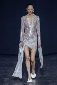 Model on a runway wearing a sheer, sequined silver dress with a plunging neckline and long cape sleeves.