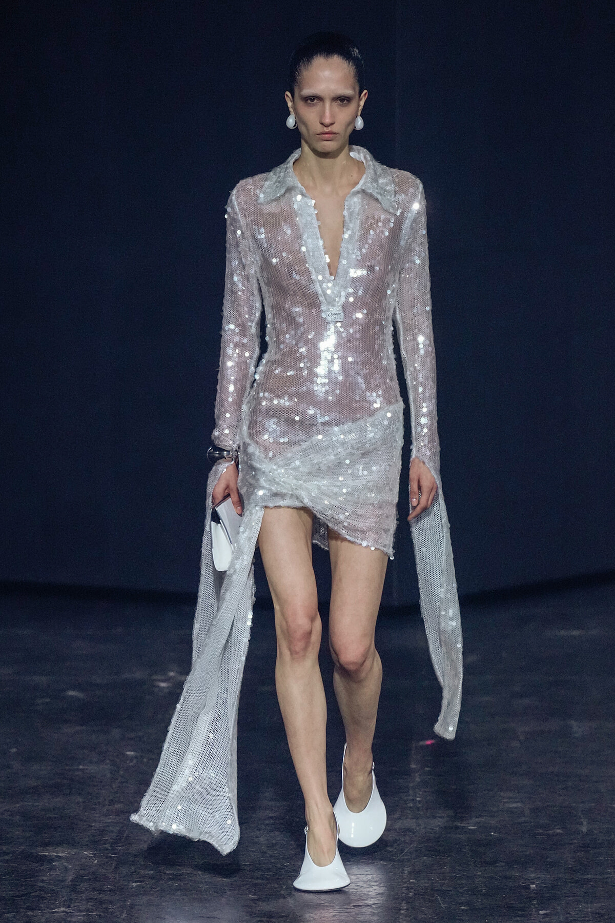 Model on a runway wearing a sheer, sequined silver dress with a plunging neckline and long cape sleeves.
