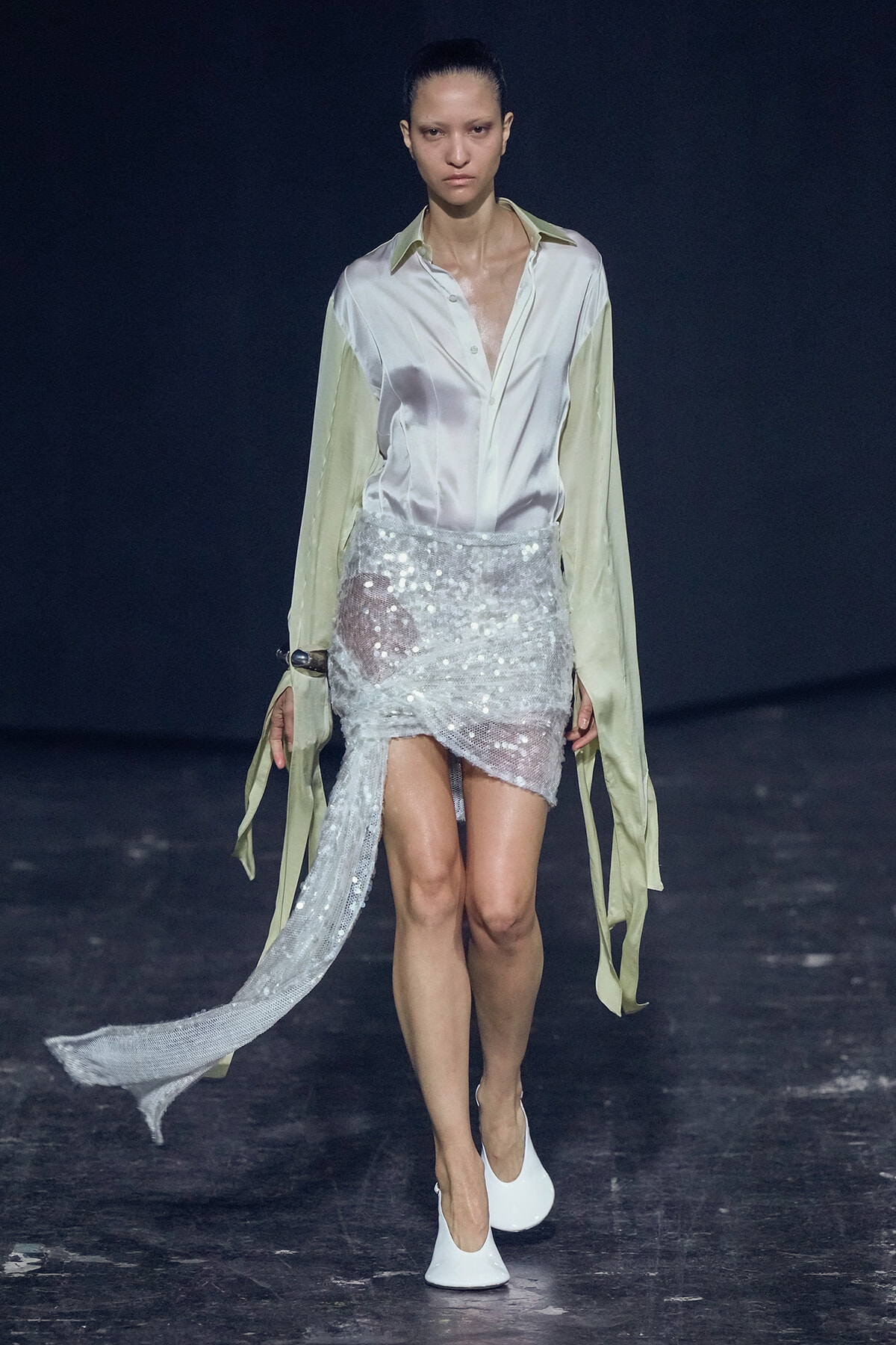 Model walks a fashion runway in a pale satin button-down shirt and a high-low silver sequin wrap skirt with white heels.