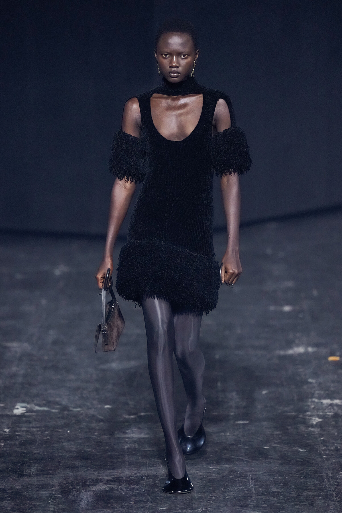 Model walking a fashion runway in a black fringe dress with open shoulders and sheer tights, carrying a small handbag