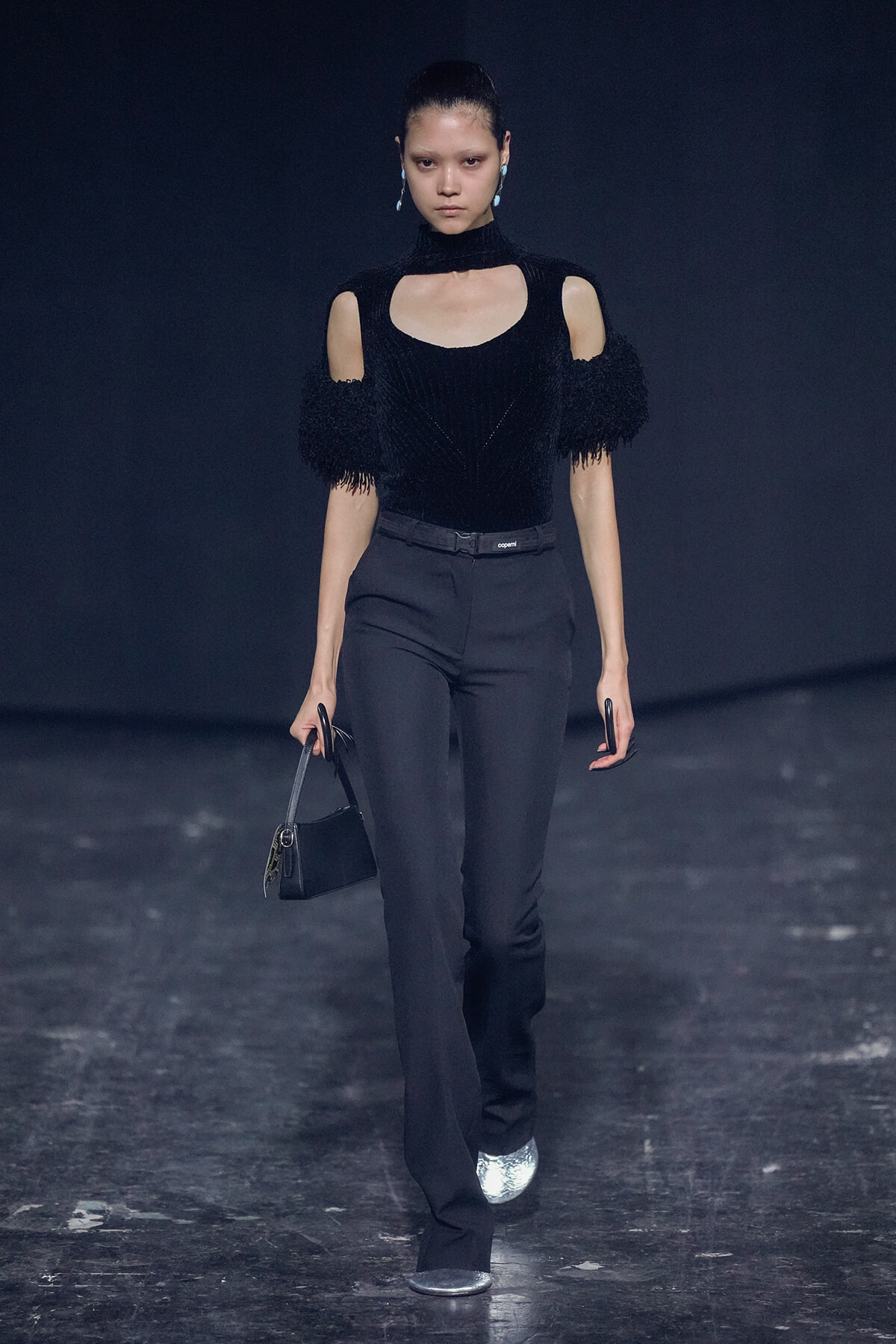 Model walks the runway in a black knit top with shoulder cutouts and fringe sleeves, paired with tailored black pants and a small handbag.