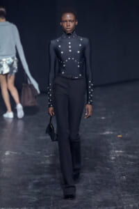 Female model on a runway wearing a black high-neck studded bodysuit with matching black trousers, carrying a small black handbag.