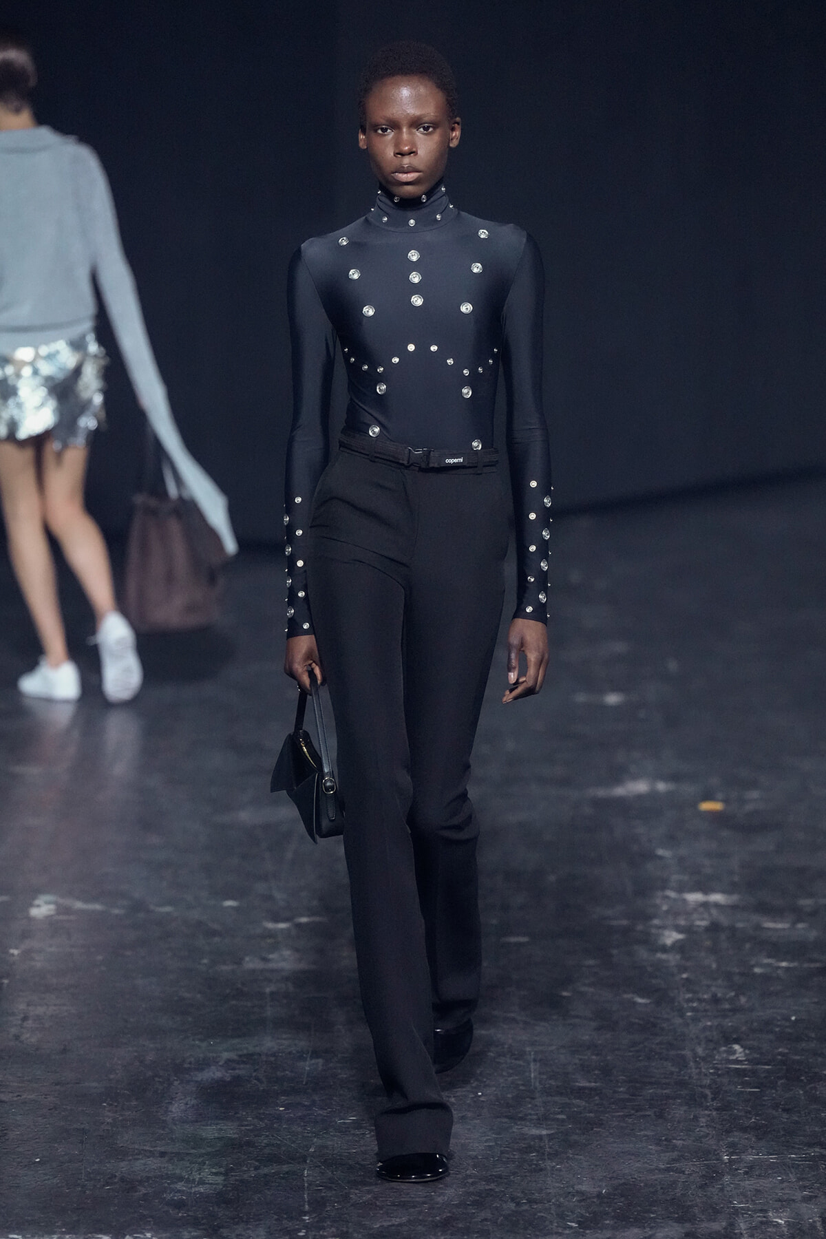 Female model on a runway wearing a black high-neck studded bodysuit with matching black trousers, carrying a small black handbag.