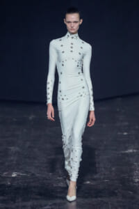 Model walking a fashion runway in a pale blue, long-sleeved, high-neck gown covered with scattered metallic studs/small discs.