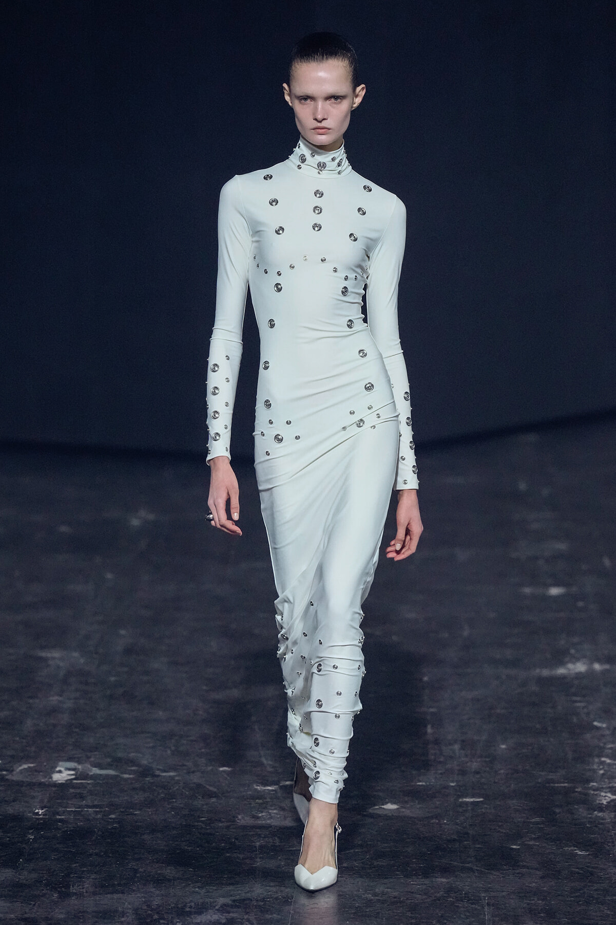 Model walking a fashion runway in a pale blue, long-sleeved, high-neck gown covered with scattered metallic studs/small discs.