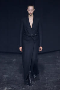 Model walking the runway in a tailored black double-breasted blazer with a wide belt, deep V-neck, and matching trousers.