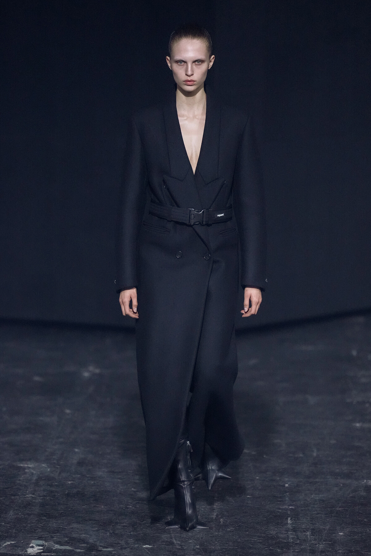 Model walking the runway in a tailored black double-breasted blazer with a wide belt, deep V-neck, and matching trousers.