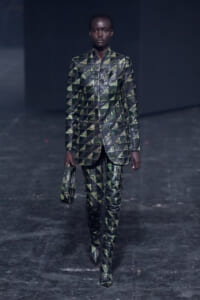 Model walks the runway in a green-and-black geometric suit with matching handbag, dark backdrop behind.