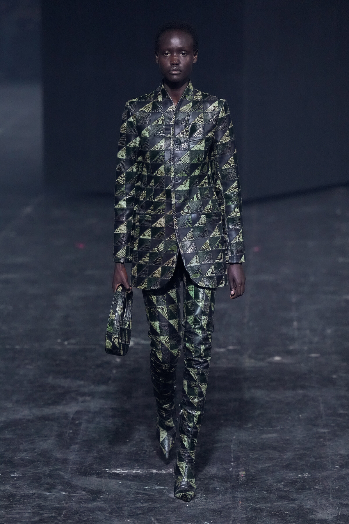 Model walks the runway in a green-and-black geometric suit with matching handbag, dark backdrop behind.