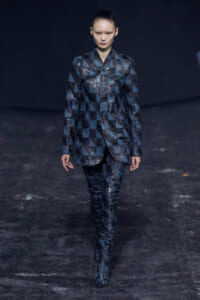 Model walks the runway in a blue geometric patchwork coat and matching pants, dark backdrop behind.