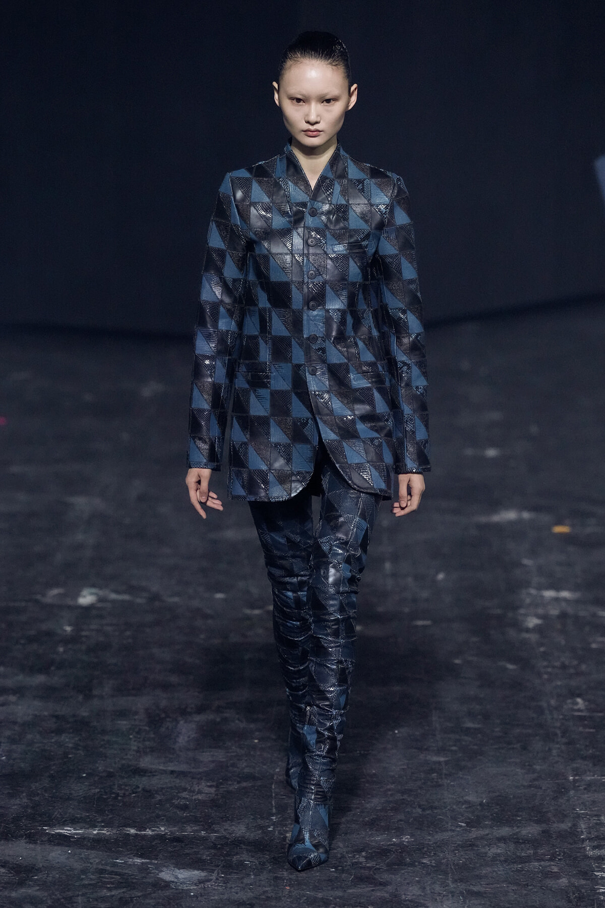 Model walks the runway in a blue geometric patchwork coat and matching pants, dark backdrop behind.
