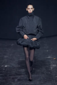 Model walking a fashion runway in an oversized dark gray jacket with a high collar and sculpted, puffed waist, paired with sheer black tights and black heels.