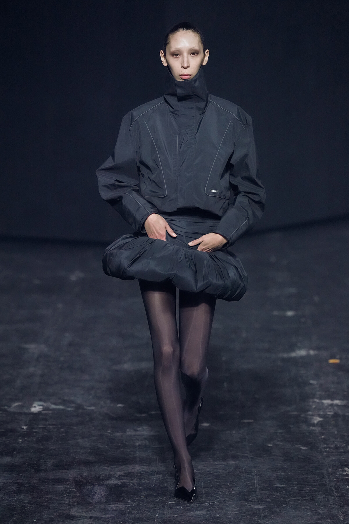 Model walking a fashion runway in an oversized dark gray jacket with a high collar and sculpted, puffed waist, paired with sheer black tights and black heels.