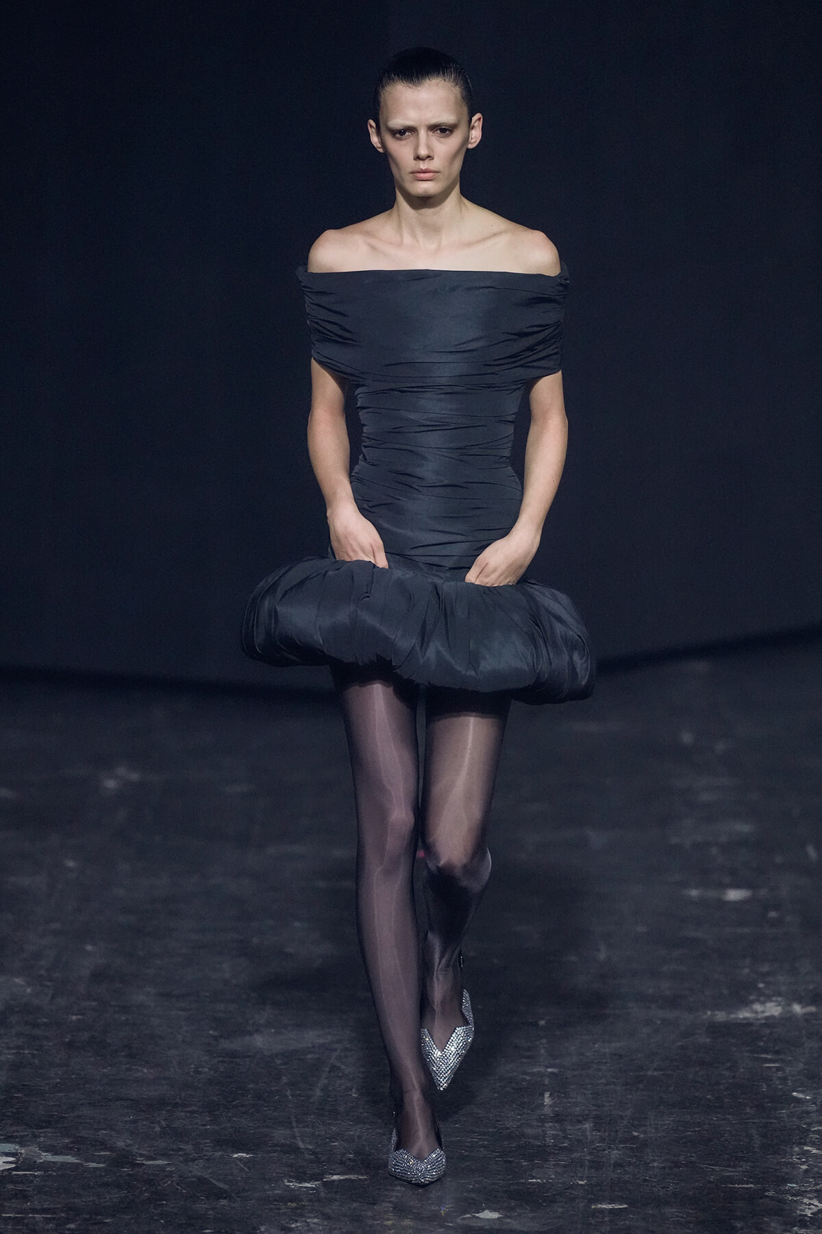 Model on a runway in a navy off-shoulder, ruched dress with a voluminous bubble hem and sparkly silver heels.