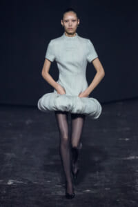 Model on a dim runway wearing a pale gray short-sleeve dress with a large circular padded hip detail, black tights, and pointed heels.