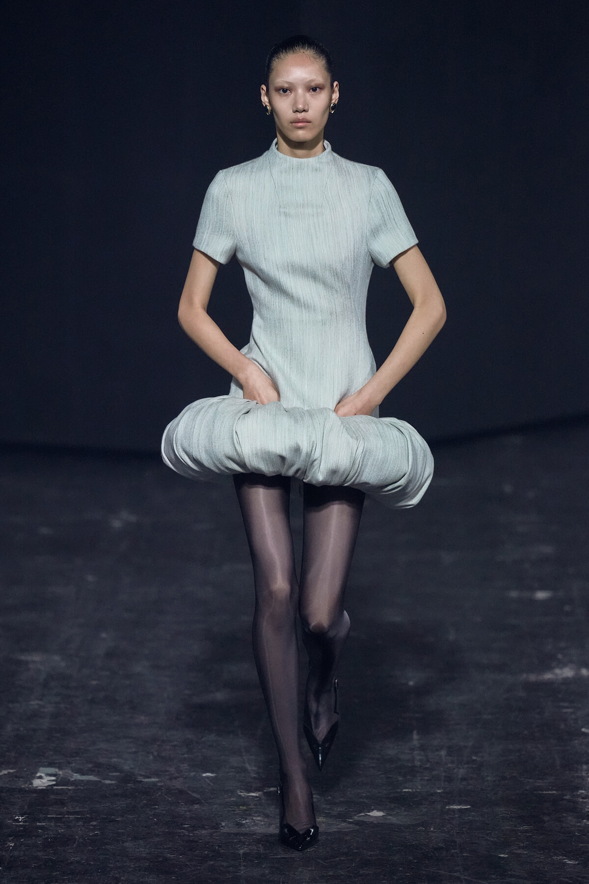 Model on a dim runway wearing a pale gray short-sleeve dress with a large circular padded hip detail, black tights, and pointed heels.